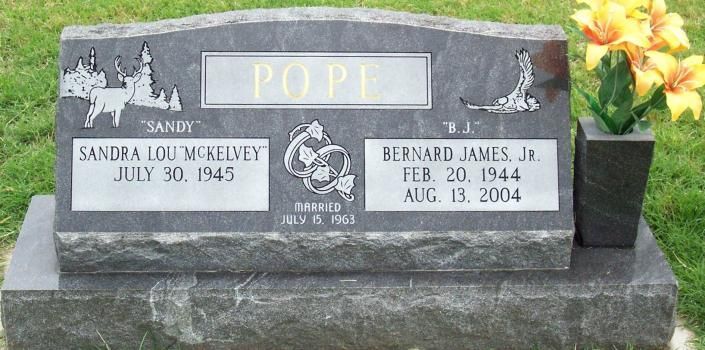 Gray memorial gravestone with “Popi” plaque and two name/date inscriptions in a grassy setting
