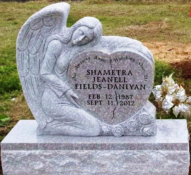 Angel-shaped gravestone with engraved name and dates, beside white flowers on a grassy cemetery plot
