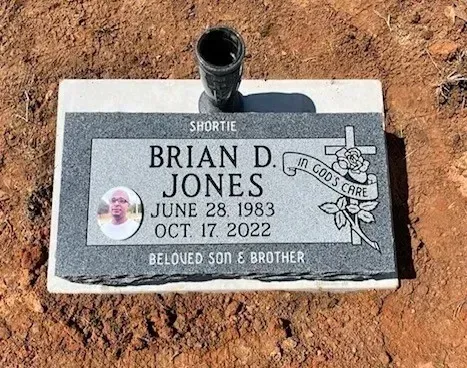 Gray memorial plaque in dirt with black vase, engraved “Brian D. Jones,” dates, and cross with flowers.