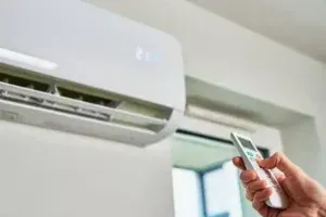 Hand Holding A Remote Pointed At A Wall Mounted Air Conditioner In A Bright Room — Alpine Refrigeration & Air Conditioning in Wollongong, NSW Hand Holding A Remote Pointed At A Wall Mounted Air Conditioner In A Bright Room — Alpine Refrigeration & Air Conditioning in Wollongong, NSW