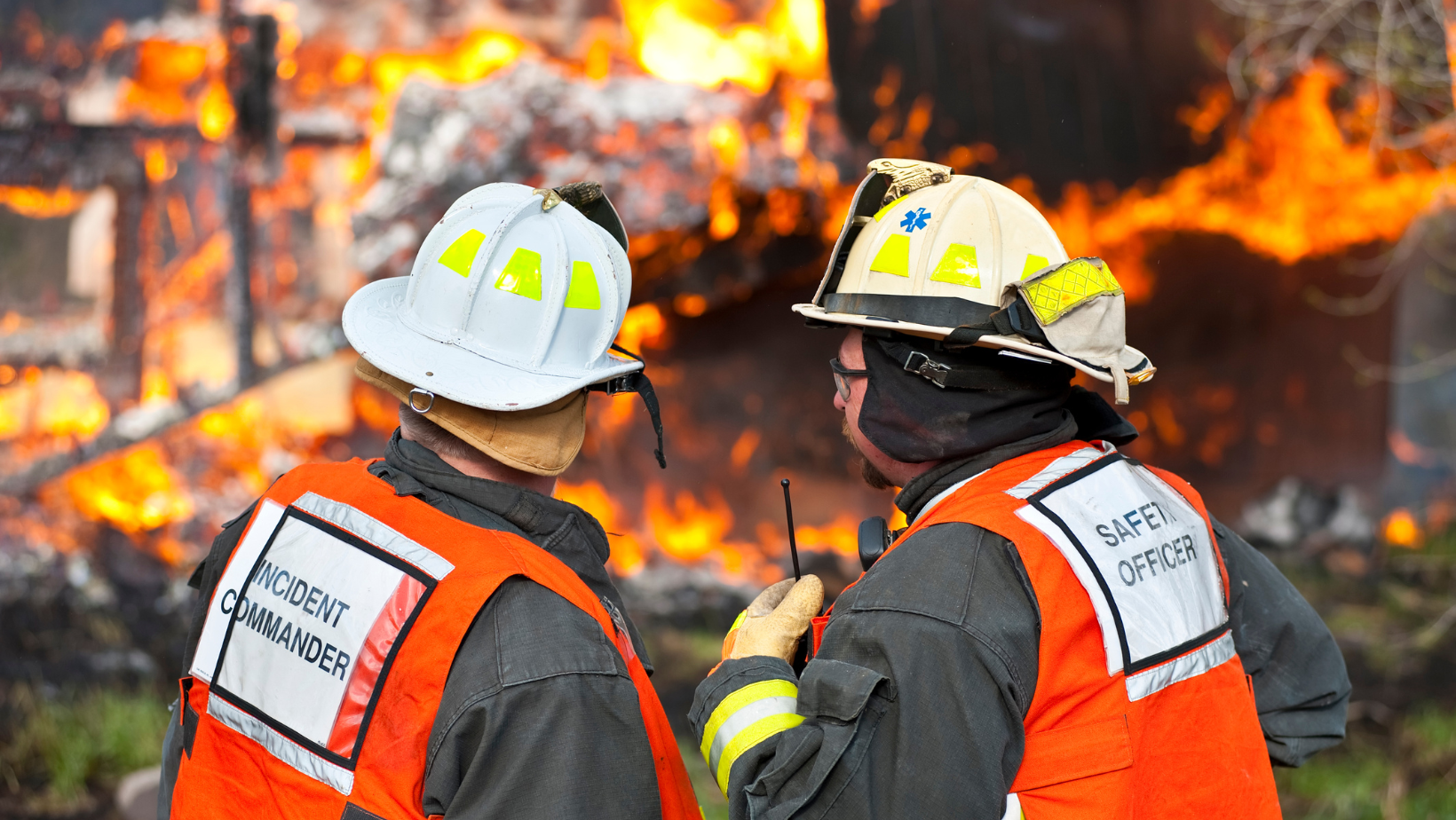 Firefighters in orange gear and white helmets face a large blaze in the background.