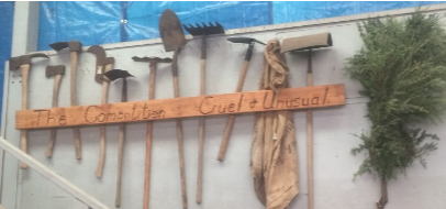 Wooden sign with metal tools and hanging cloth mounted on a wall, beside greenery.