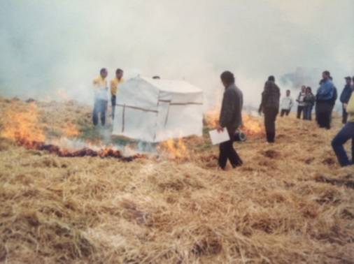 People standing near a burning field beside a white tarp shelter in a smoky outdoor area