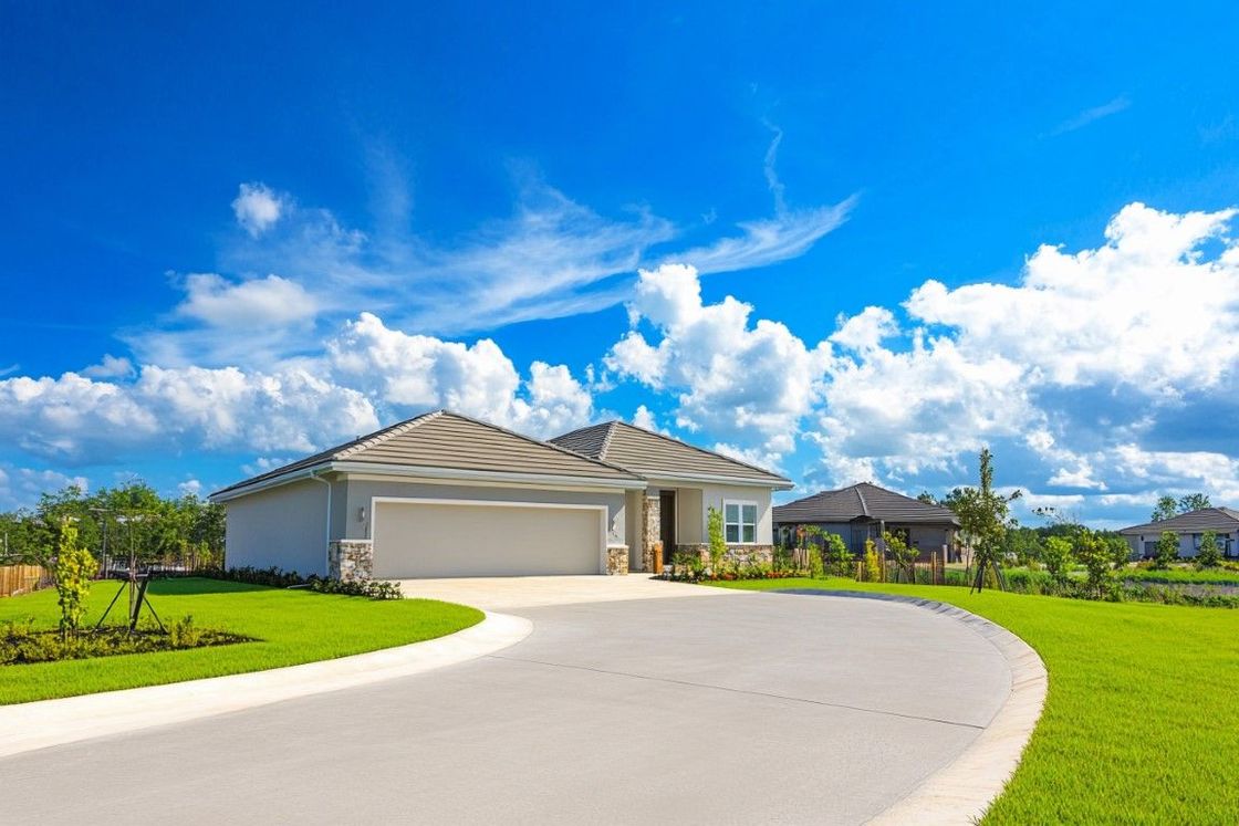 A light-colored house with a curved driveway and green lawn under a bright blue sky with clouds.