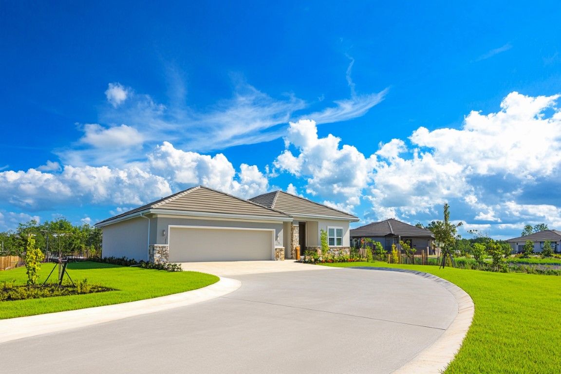 A light-colored house with a curved driveway and green lawn under a bright blue sky with clouds.