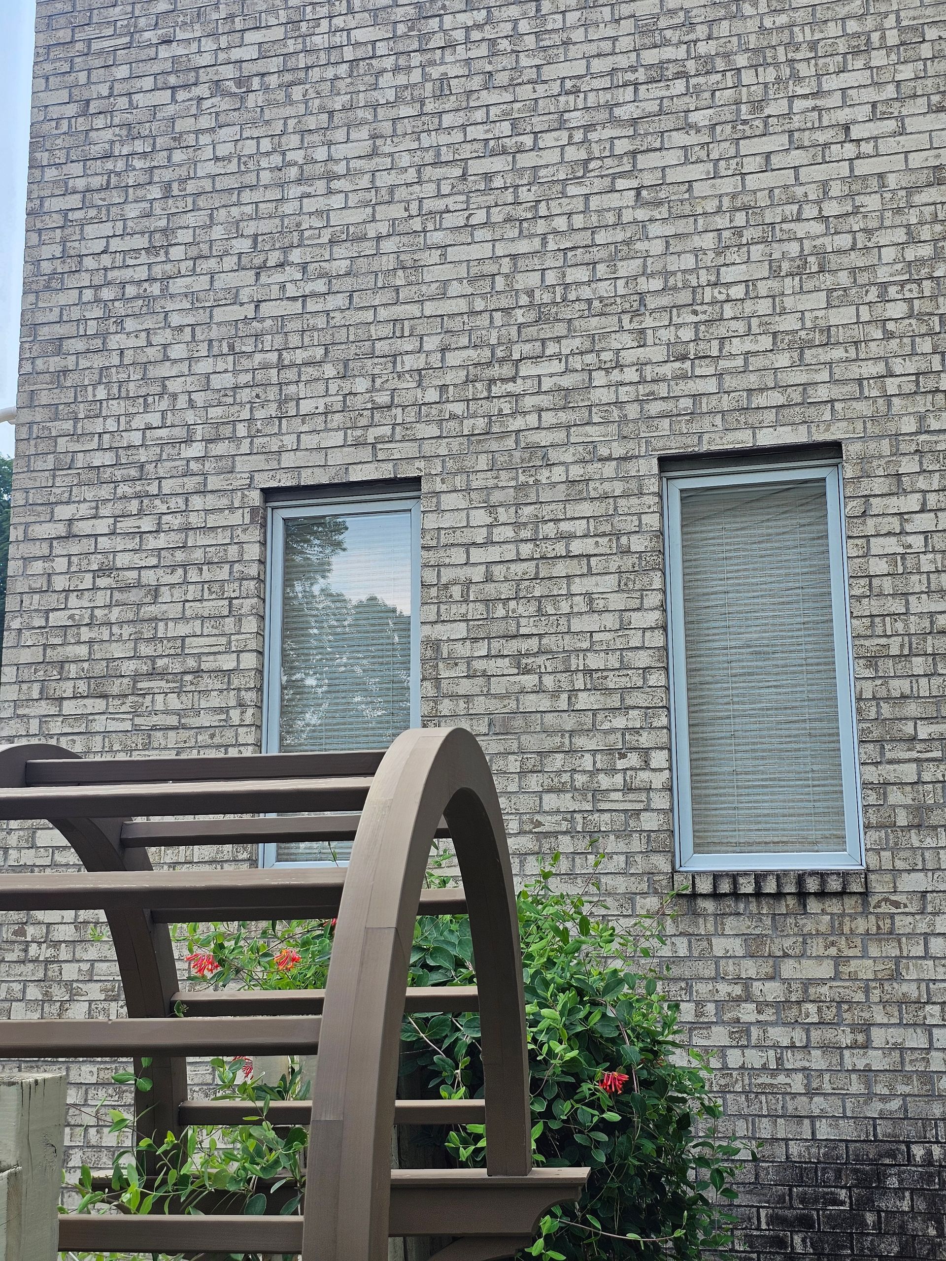 Brick building with two windows, a wooden staircase, and green foliage.
