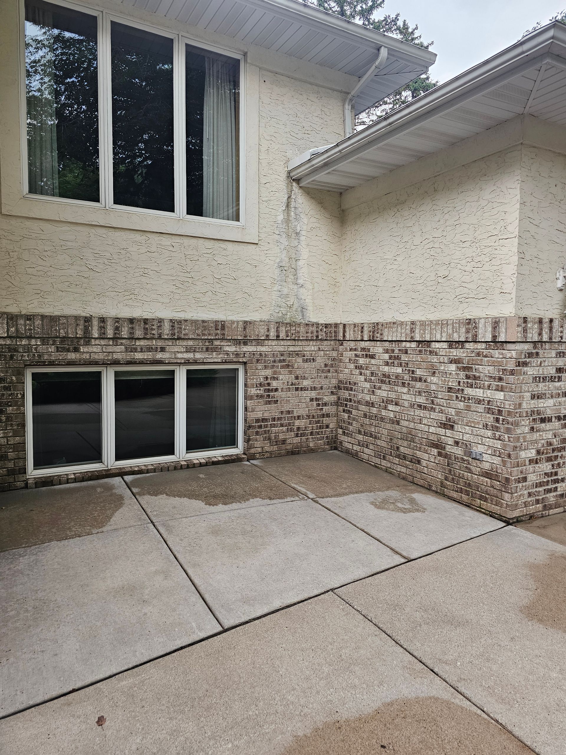 Exterior of a brick and stucco house with large windows, concrete patio, and wet ground.