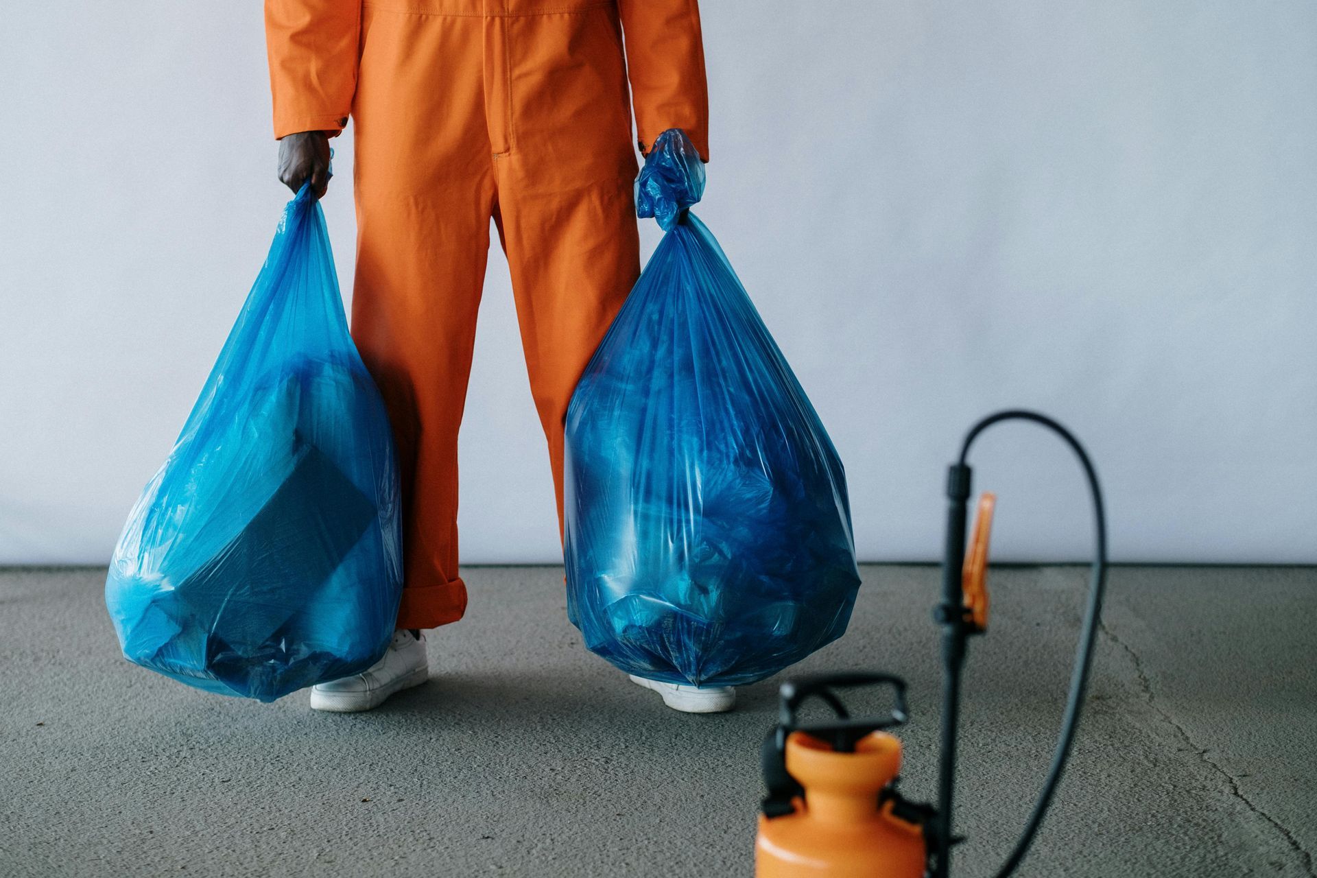 Person in orange jumpsuit holds two blue trash bags, indoors with a sprayer.