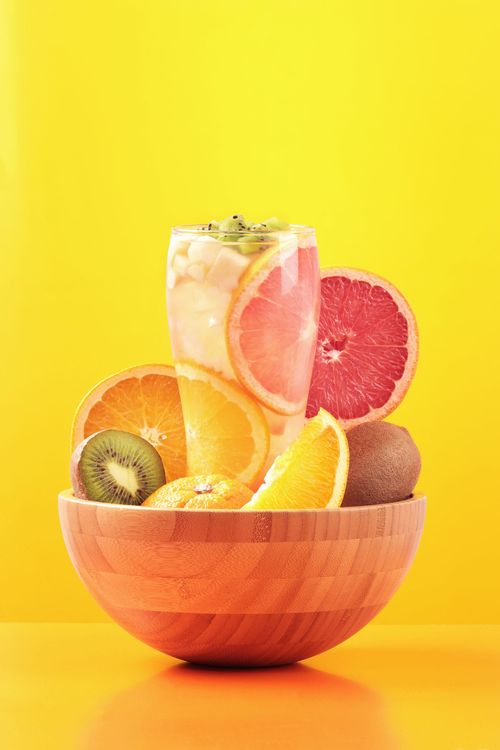 Wooden bowl of assorted fresh fruit on a bright yellow background with a glass of fruit drink behind it