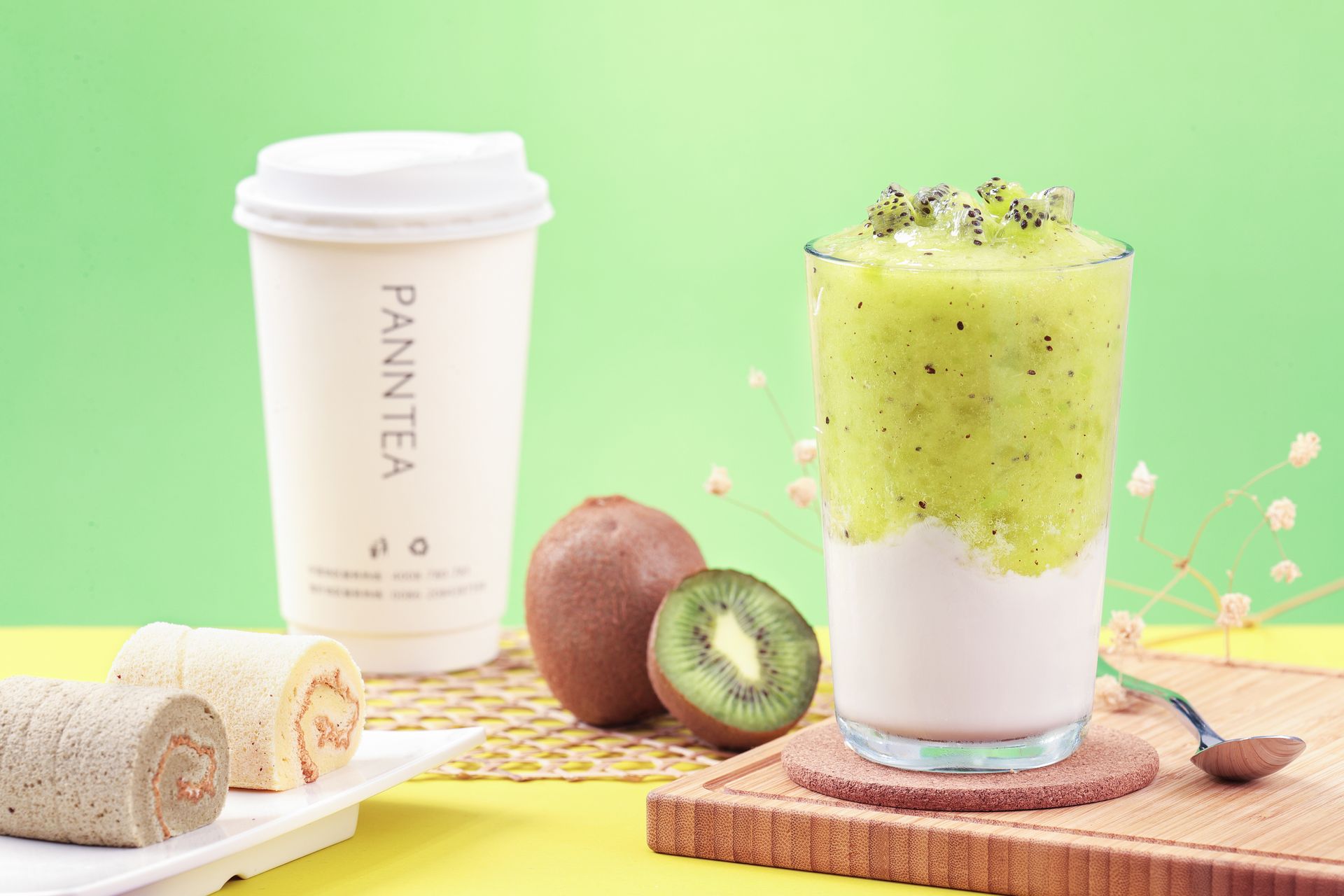 Two kiwi smoothies on a pastel table with sliced fruit, wrapped rolls, and a takeaway cup against a green backdrop