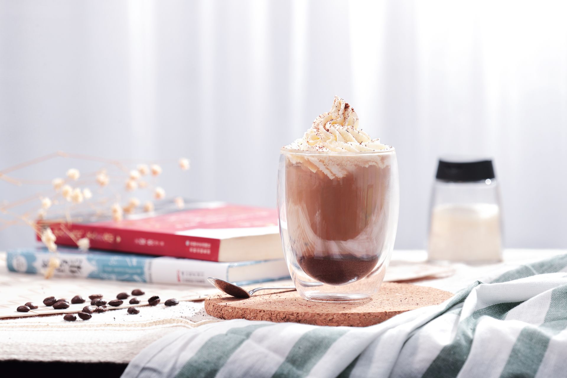 Iced coffee with whipped cream beside stacked books and scattered coffee beans on a table