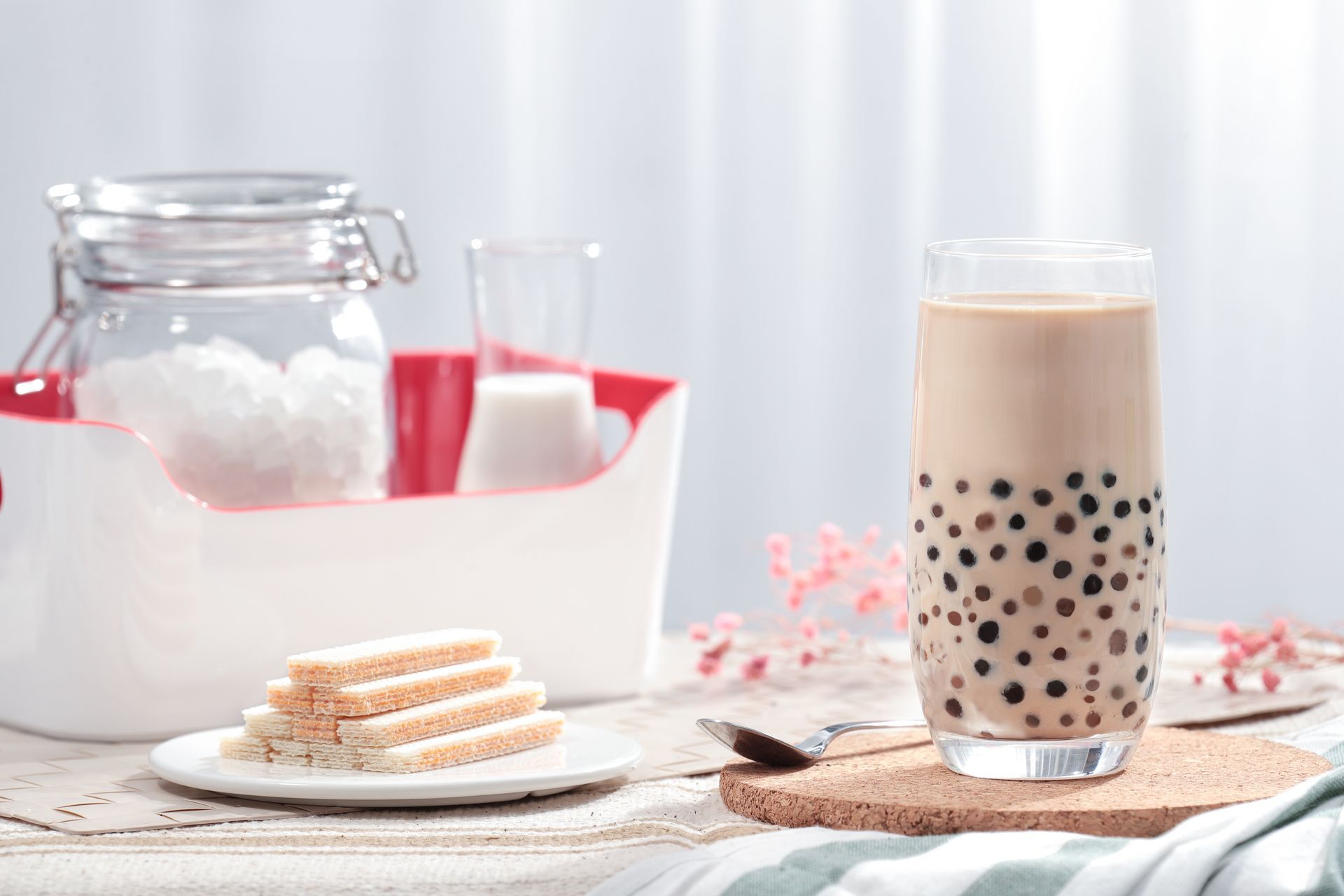 Glass of bubble tea with tapioca pearls beside a plate of pastries and a white jar on a bright table