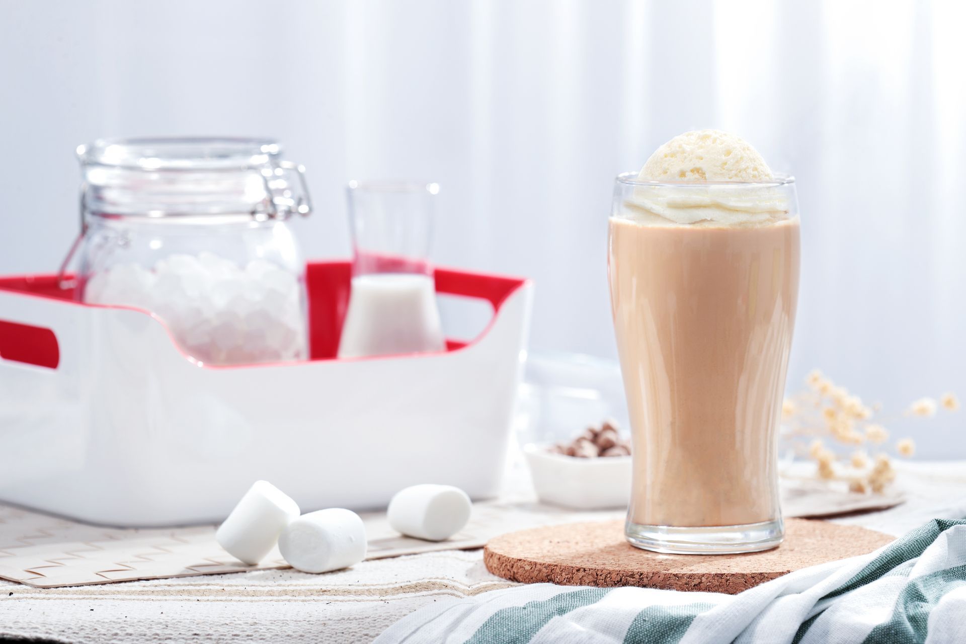 Glass of iced coffee with whipped cream beside a white cooler, ice, and marshmallows on a bright table