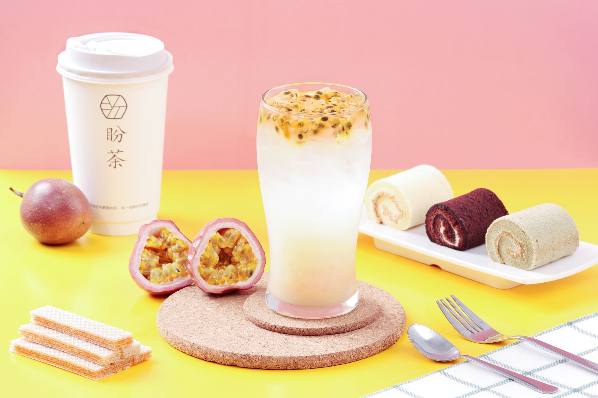 Pink dessert-and-drink display with two takeaway cups, fruit, pastries, and a fork on a yellow table