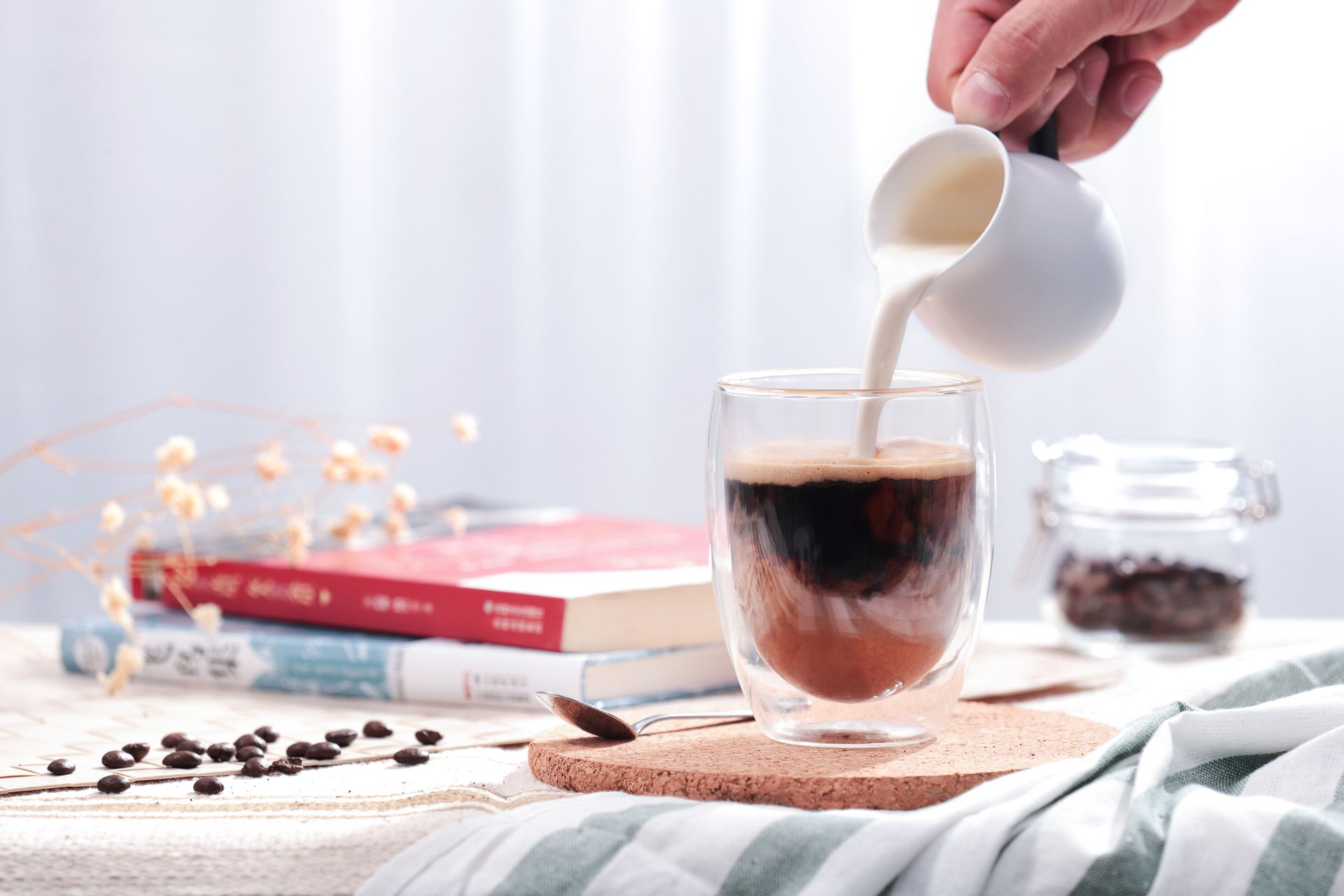Hand pouring milk into a glass of iced coffee on a bright table with coffee beans and a book