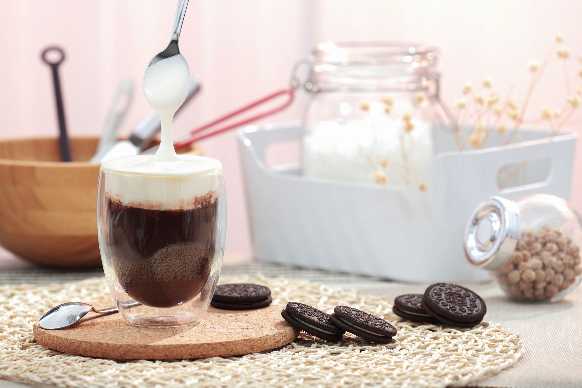 Iced coffee in a glass with milk being poured, beside cookies on a woven mat in a bright kitchen scene