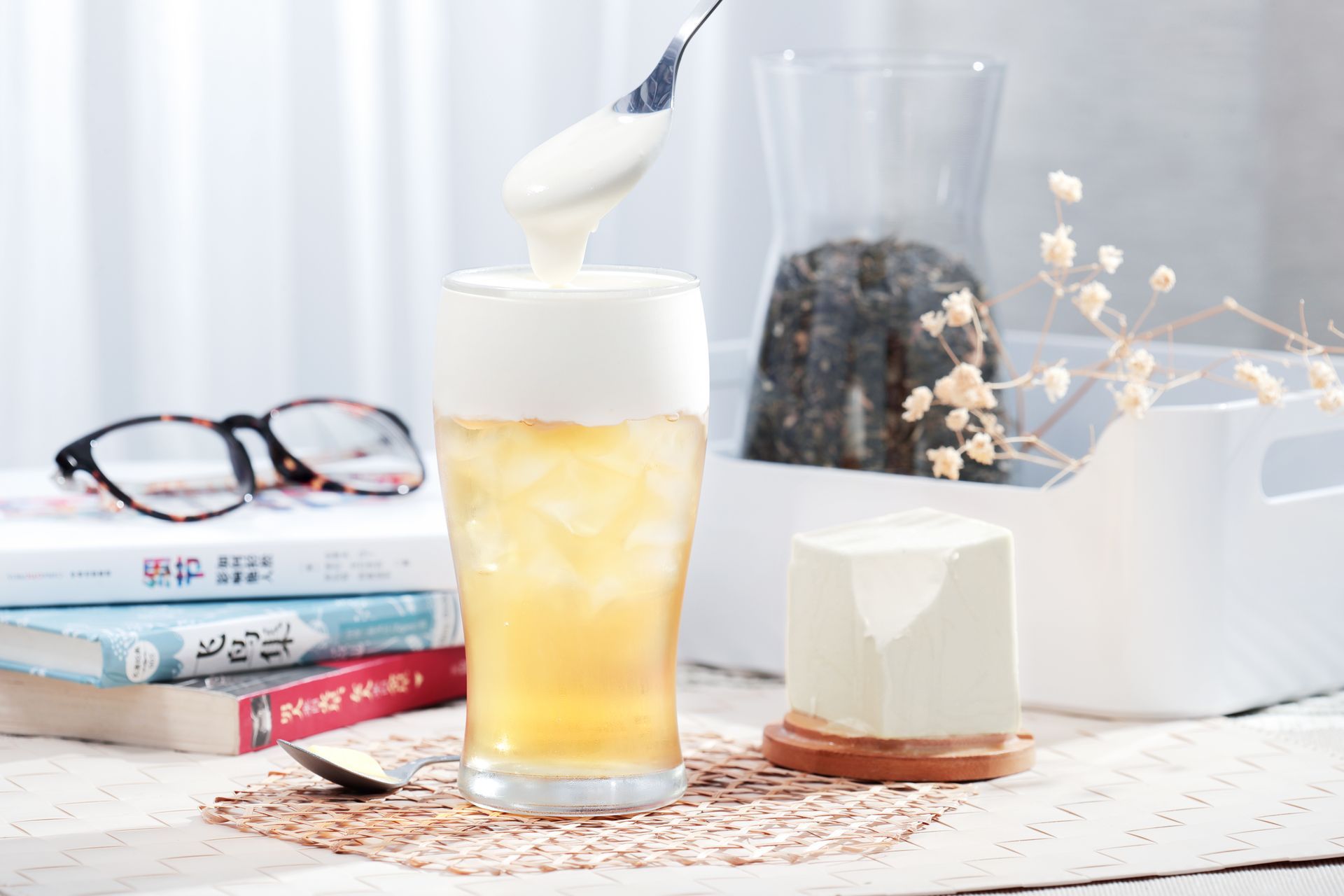 Glass of iced drink with a spoon beside books, glasses, and a candle on a bright table
