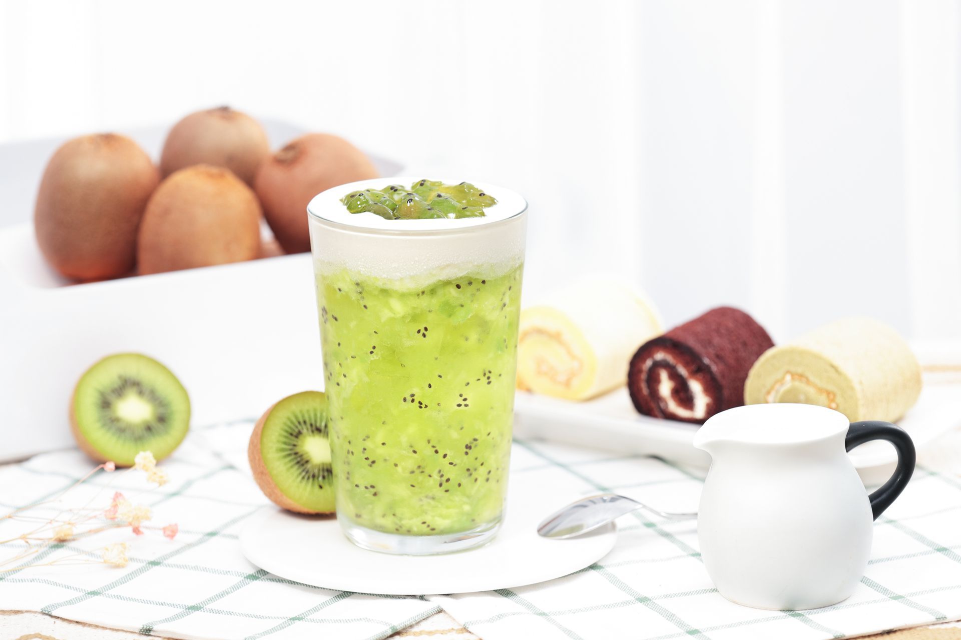 Glass of bright green kiwi smoothie with sliced kiwis, pastries, and eggs on a white table