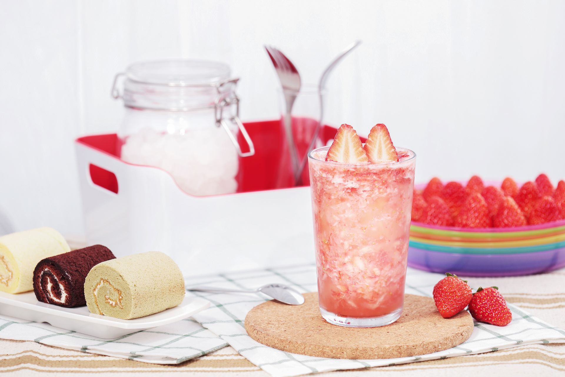 Dessert drink with strawberries on a woven mat beside a jar, fruit bowl, and rolled cakes on a white table