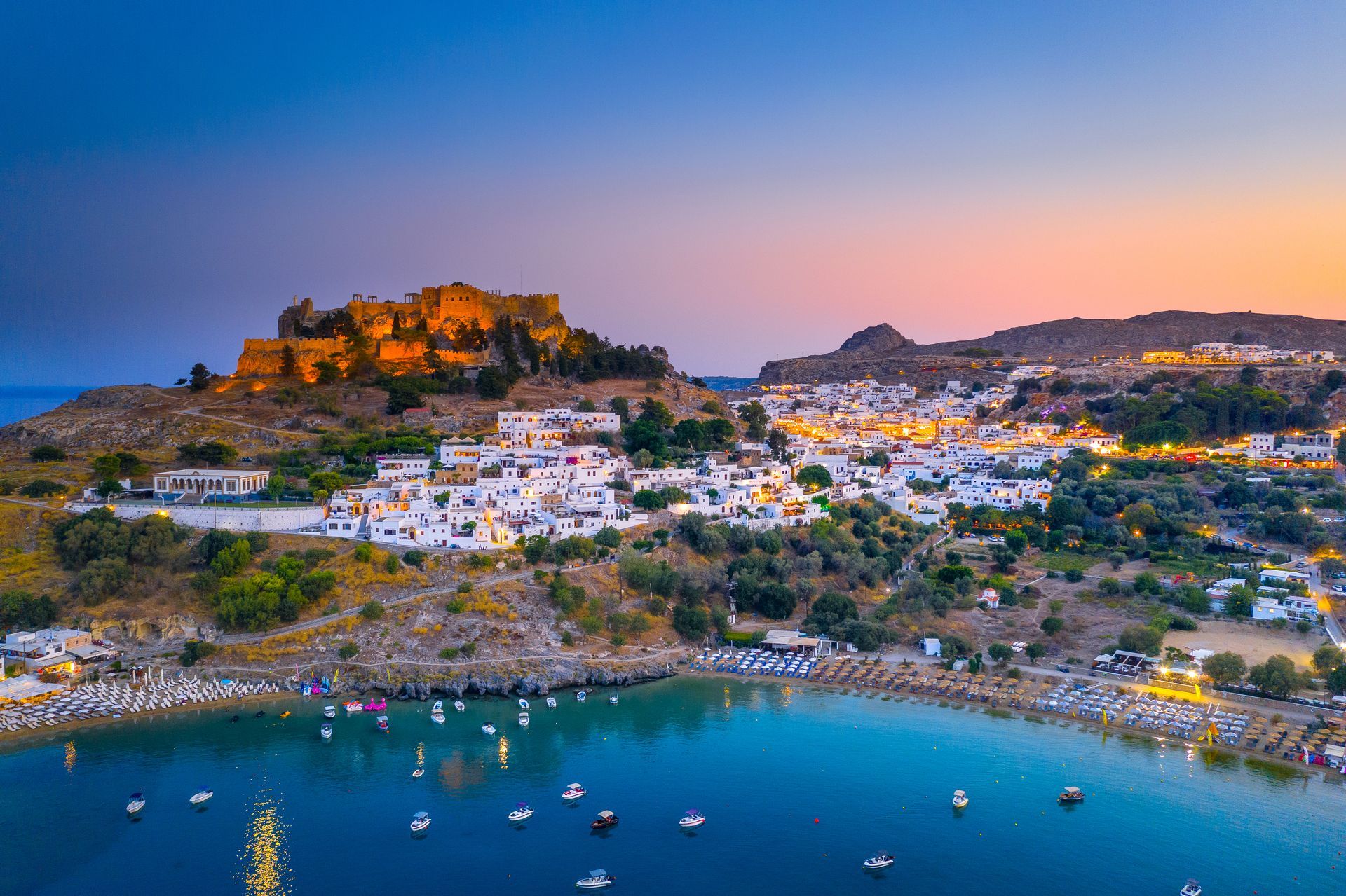 Coastal town at dusk with white buildings, lit hilltop ruins, and boats in a calm blue bay