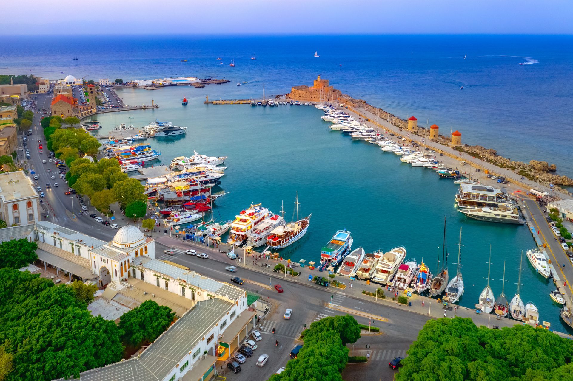 Aerial view of a marina with colorful boats, a waterfront road, and blue sea under a clear sky