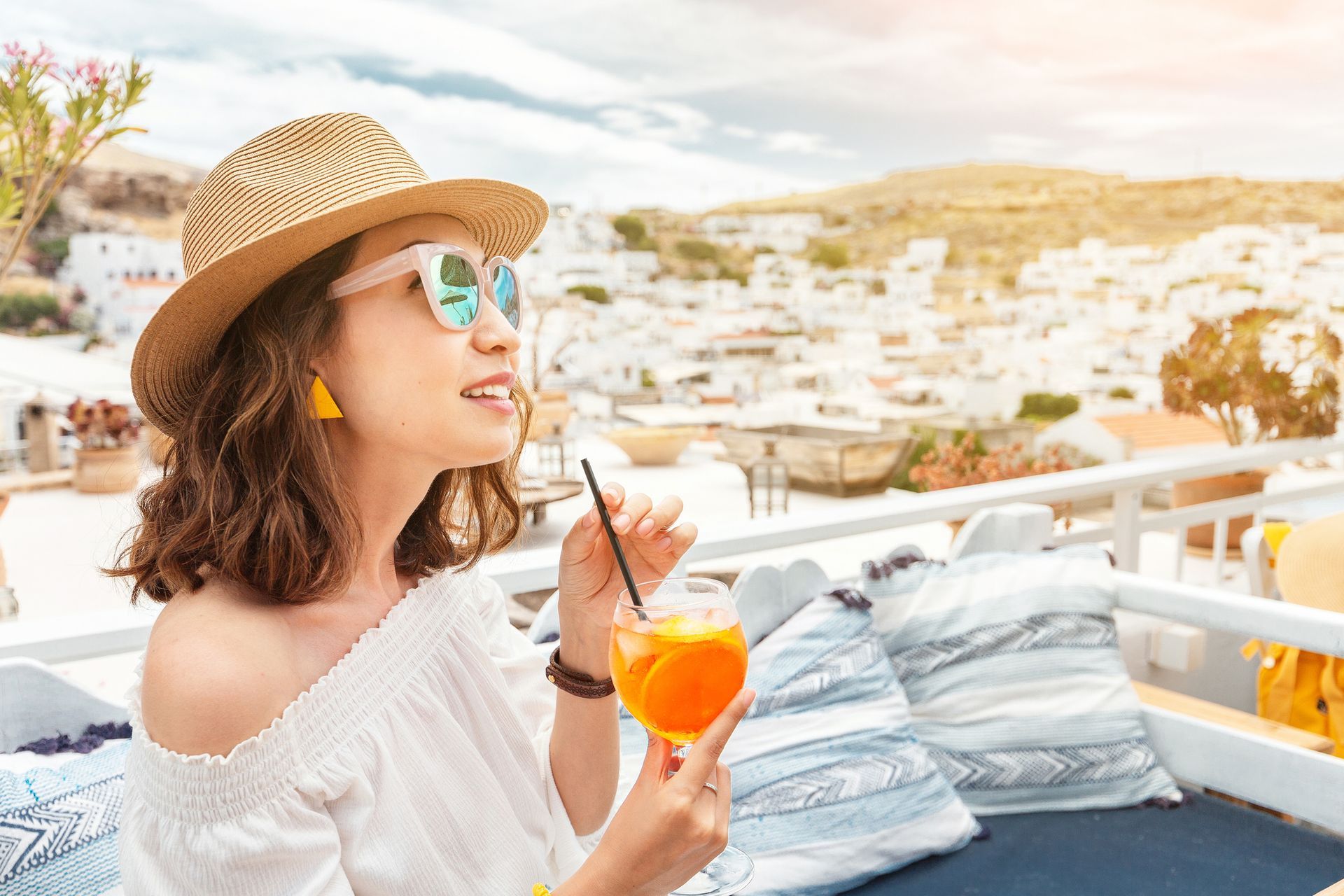 Woman in straw hat sipping an orange drink on a sunlit rooftop terrace with white buildings in the background