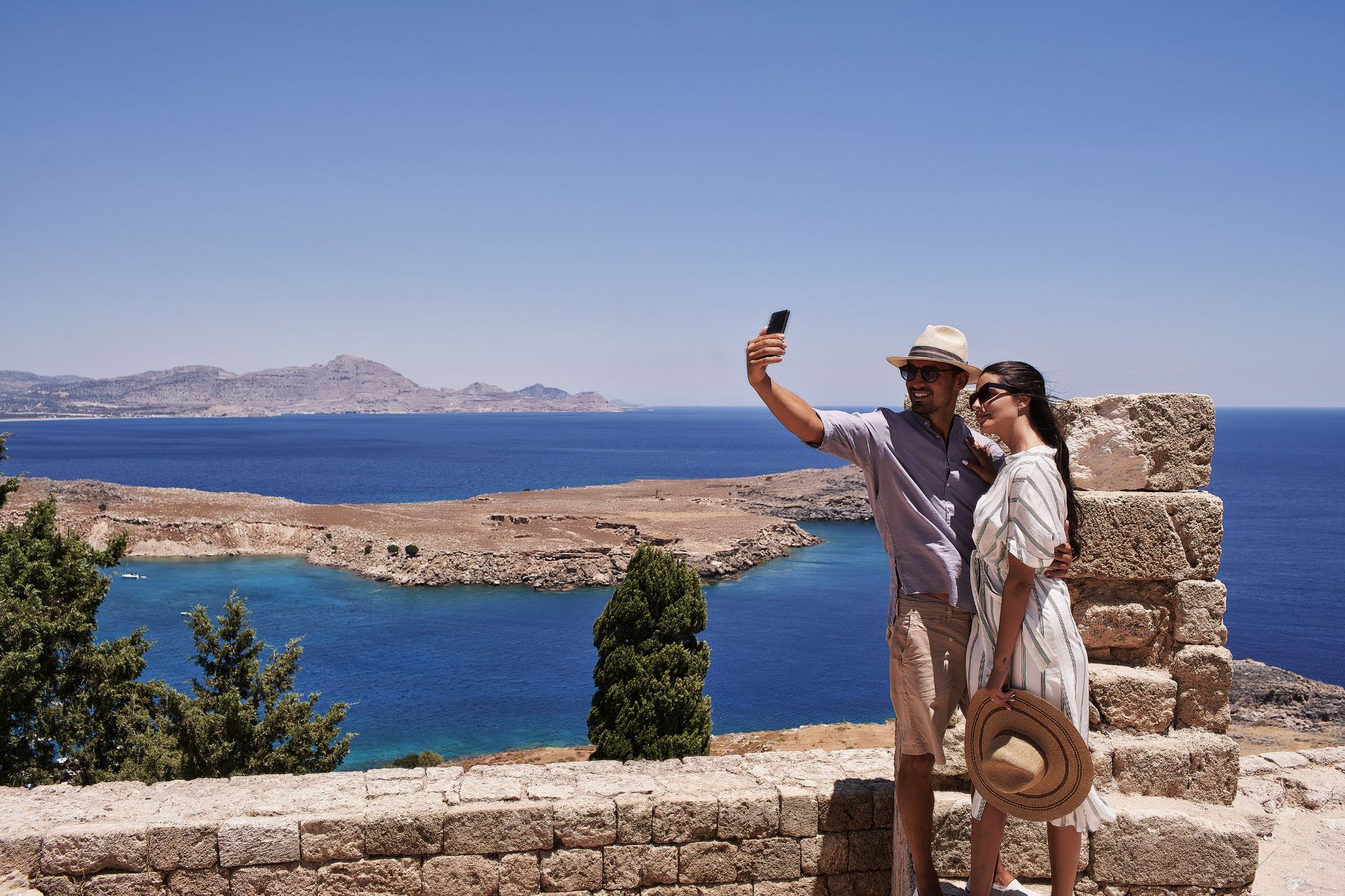 Two people taking a selfie by a stone wall with blue sea and rocky coastline in the background