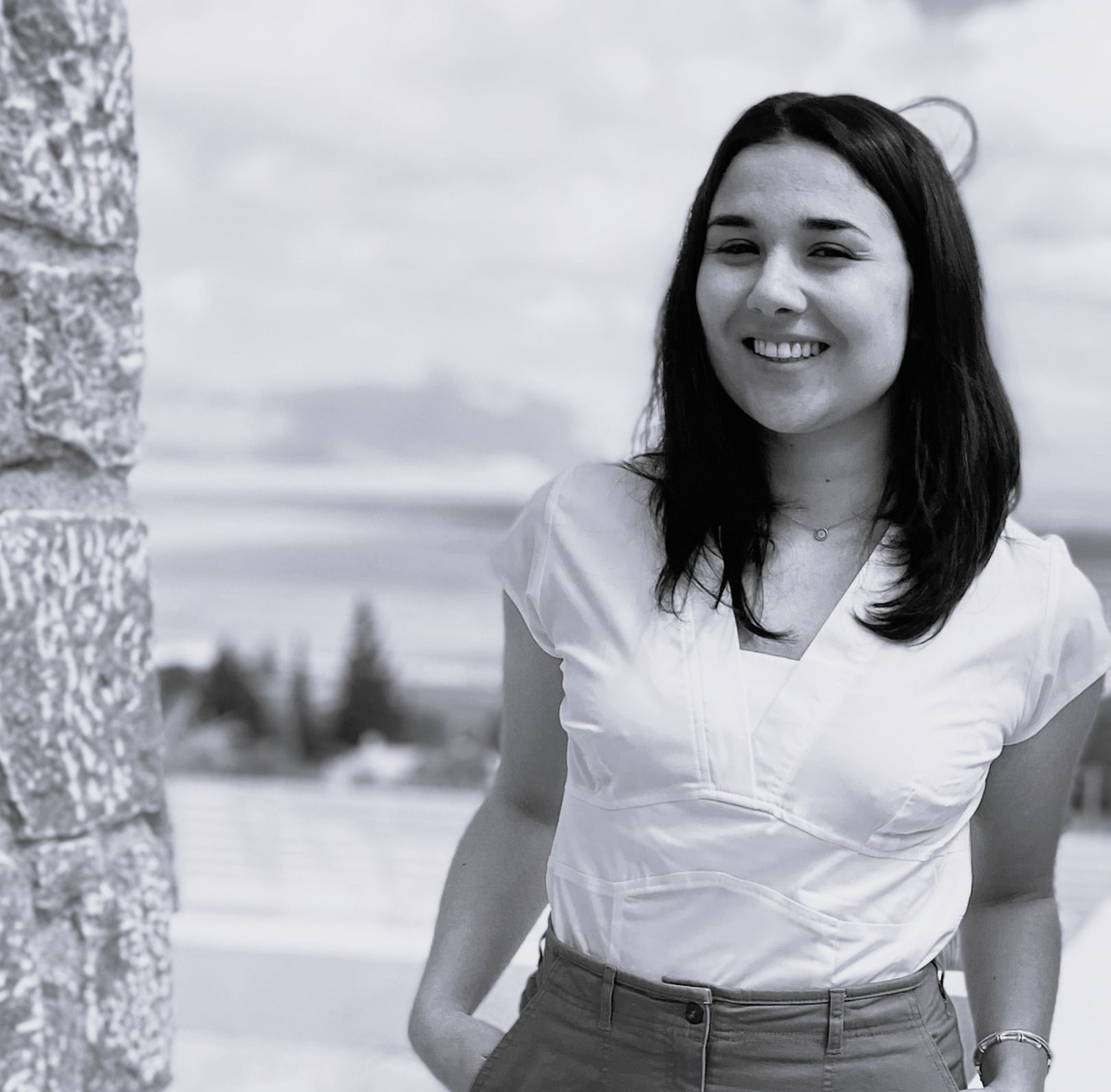 Woman smiling outdoors beside a stone wall, with mountains in the background.