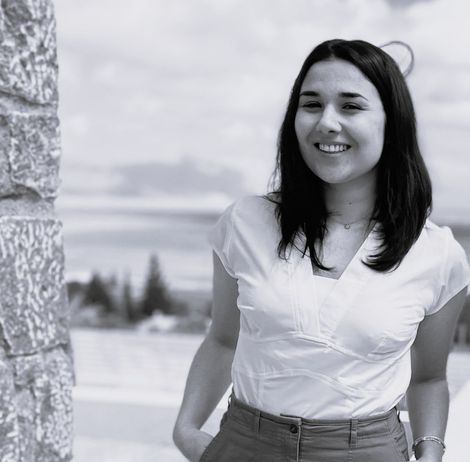Smiling woman in a white blouse standing outdoors beside a stone wall with mountains in the background