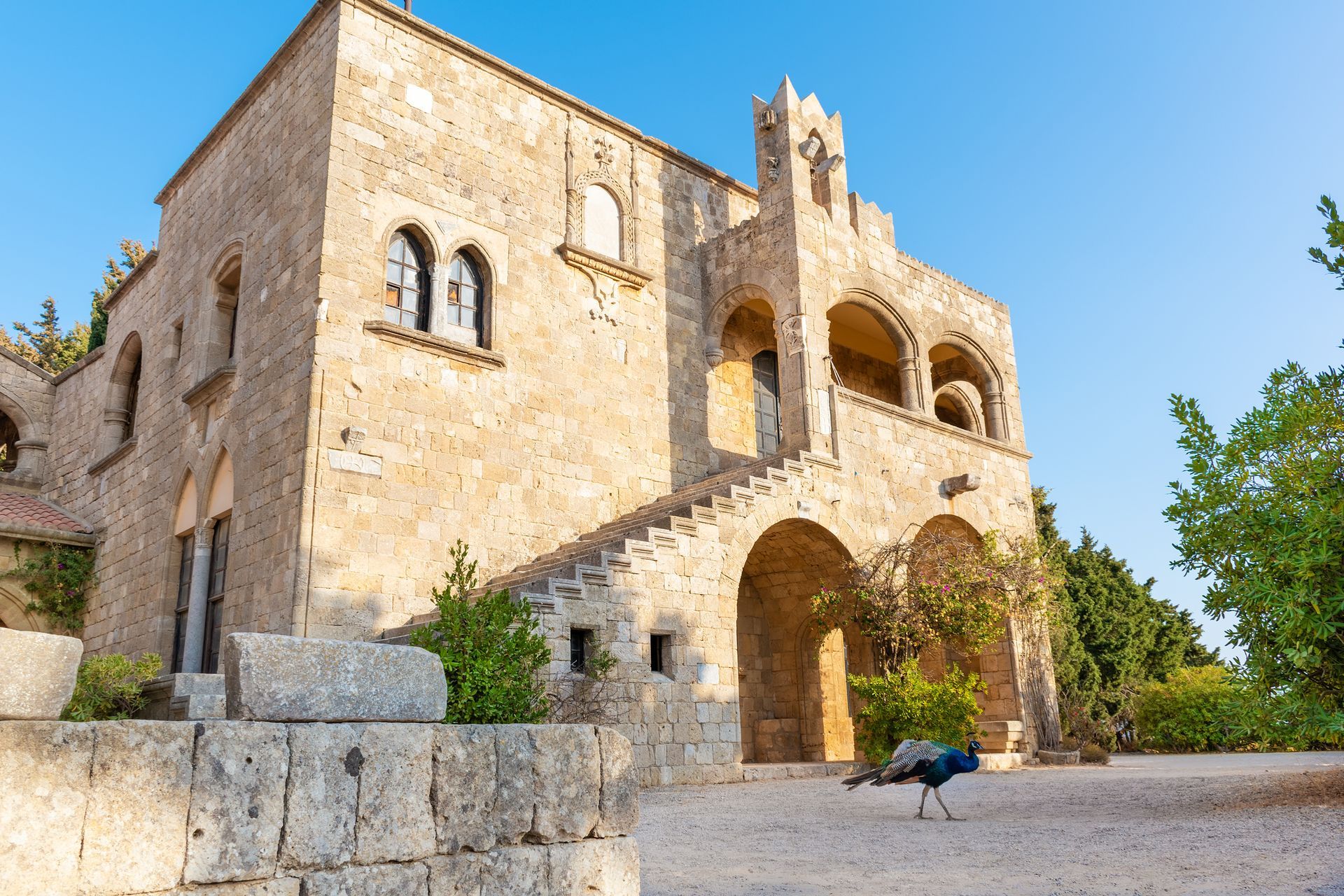 Stone medieval building with arched entrance and tower, viewed from a gravel courtyard under a blue sky.