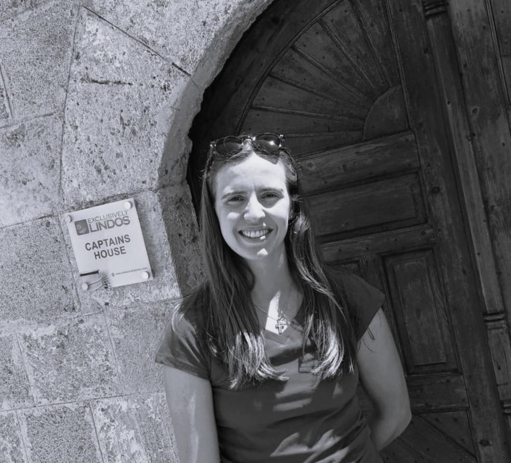 Smiling woman standing by a stone archway and dark wooden door, with a small sign on the wall.
