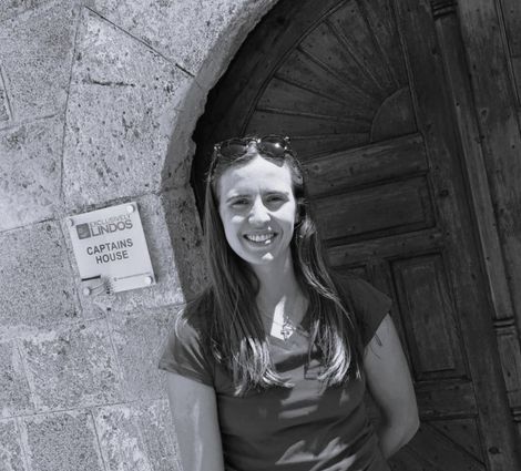 Smiling woman standing by a stone archway and wooden door, with a small sign on the wall nearby.