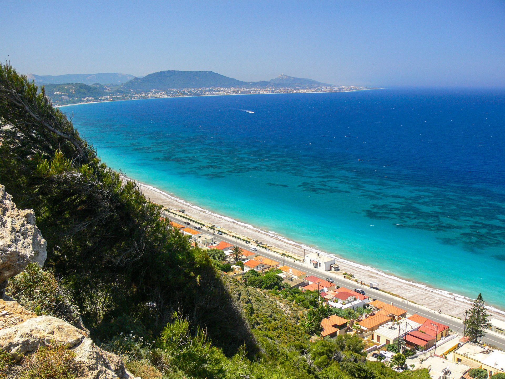 Coastal view of turquoise sea, sandy beach, and hillside with red-roofed buildings under a clear blue sky
