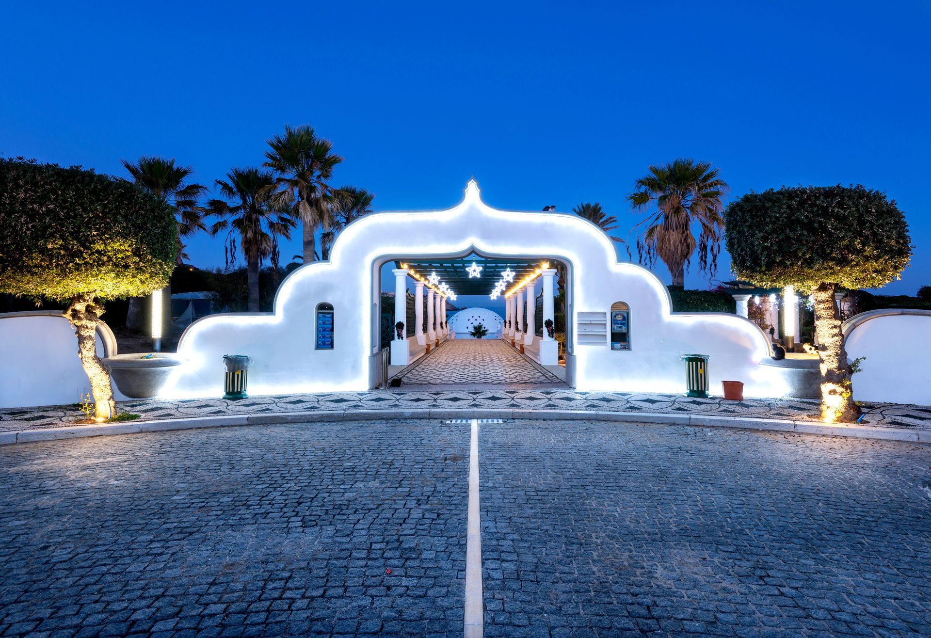 Illuminated white arched entrance at dusk, lined by palm trees and cobblestone driveway.