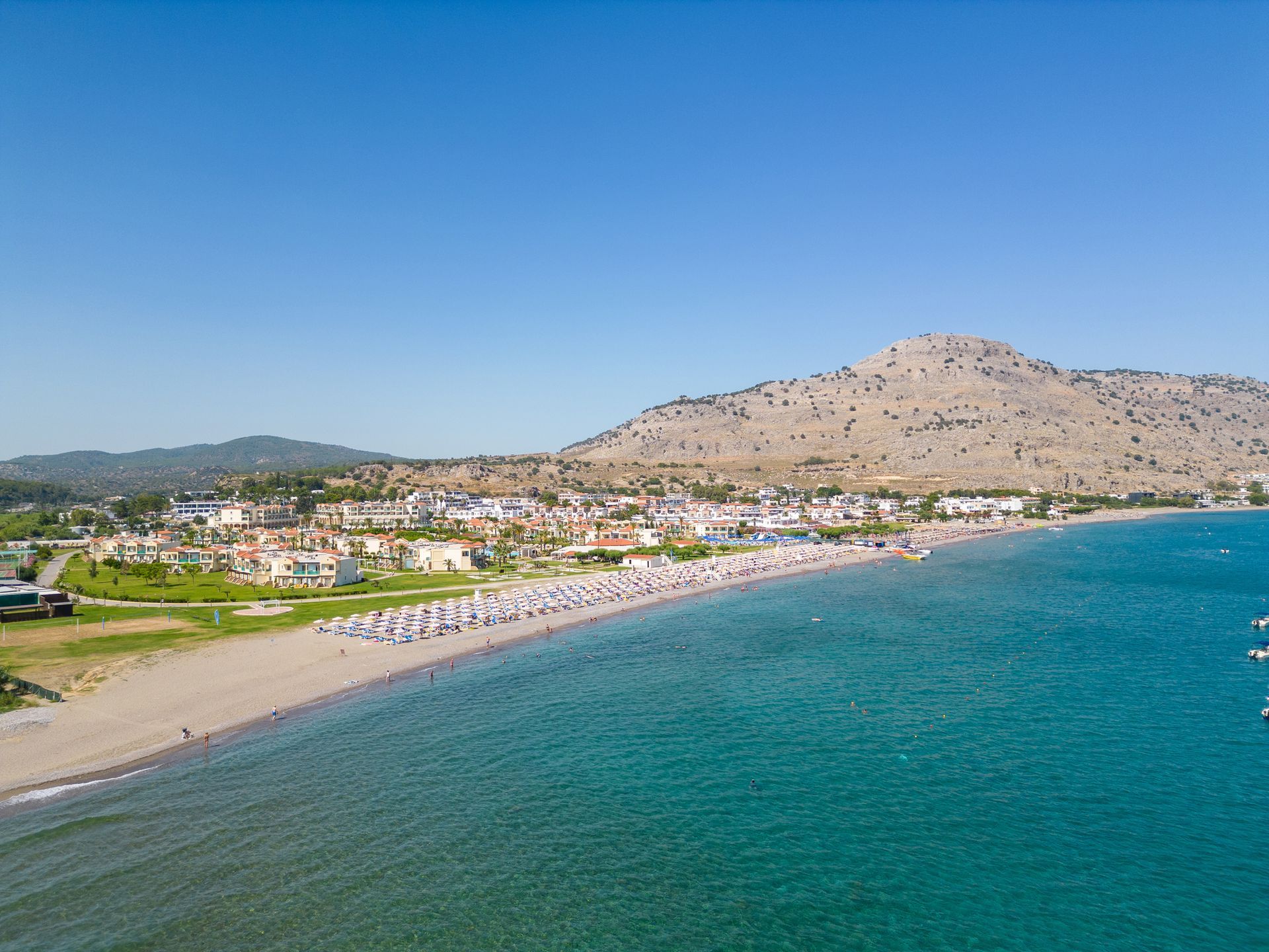 Coastal beach town with turquoise water, sandy shore, and hillside homes under a clear blue sky