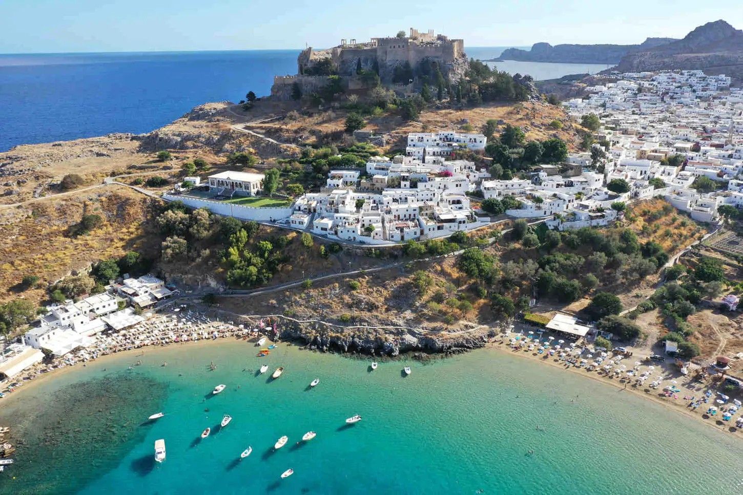 Aerial view of a coastal town with a hilltop castle, white buildings, and boats in a turquoise bay