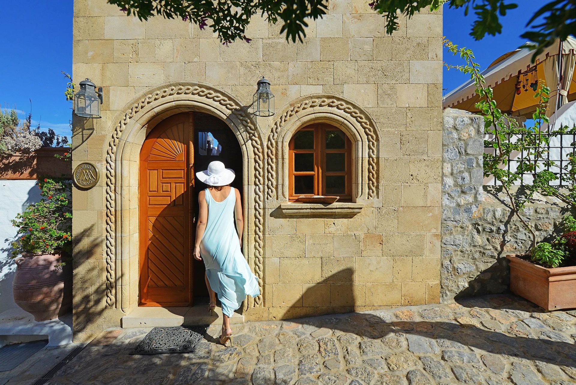 Woman in a white dress and hat entering a stone cottage with an arched wooden door.