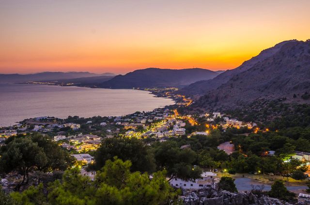 Sunset over a coastal bay with mountains, a town below, and purple-orange sky