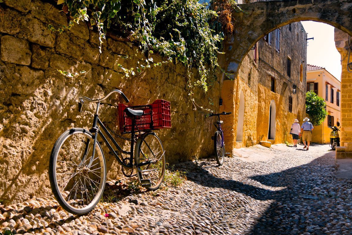 Bicycle with a red basket parked on a cobblestone alley beside stone walls in warm sunlight