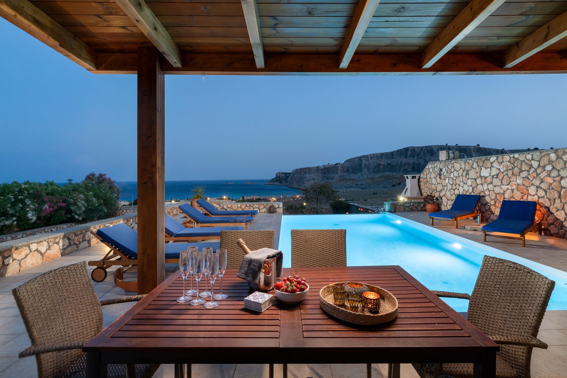 Seaside patio with dining table, infinity pool, and ocean view at sunset.