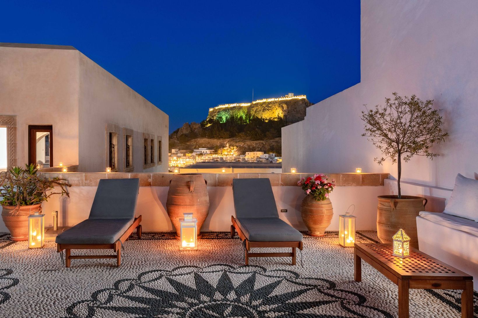 Courtyard lounge with lit fire pits, lounge chairs, and a lit hillside terrace at dusk