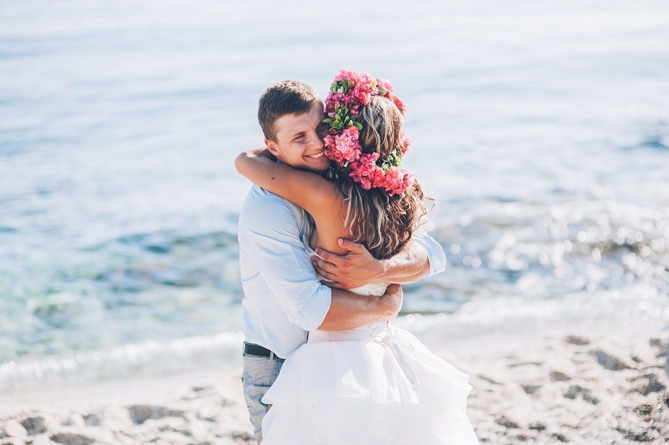 Couple embracing on a bright beach, the woman in a white dress with a flower crown.