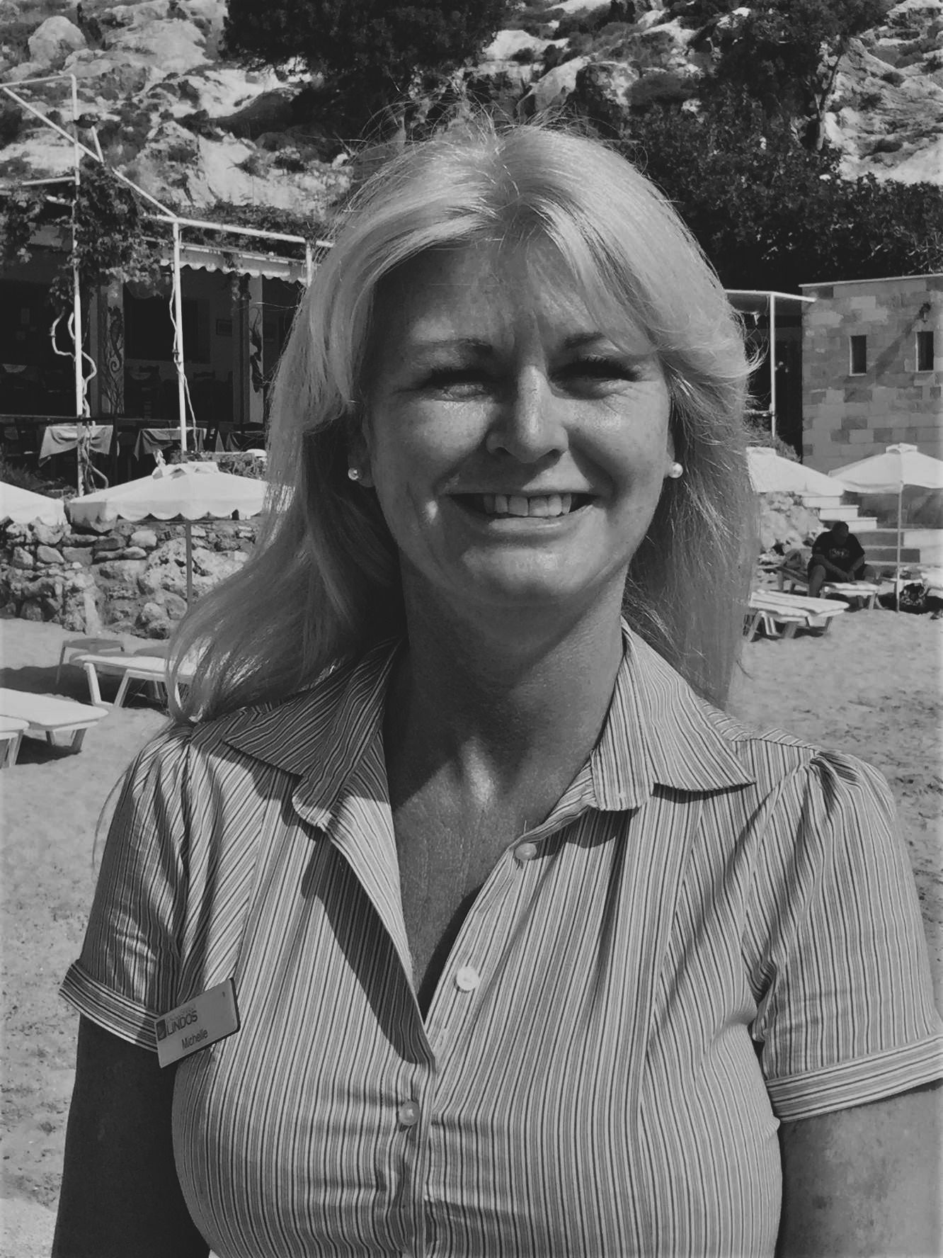 Smiling woman outdoors in a striped shirt, with lounge chairs and a resort pool behind her