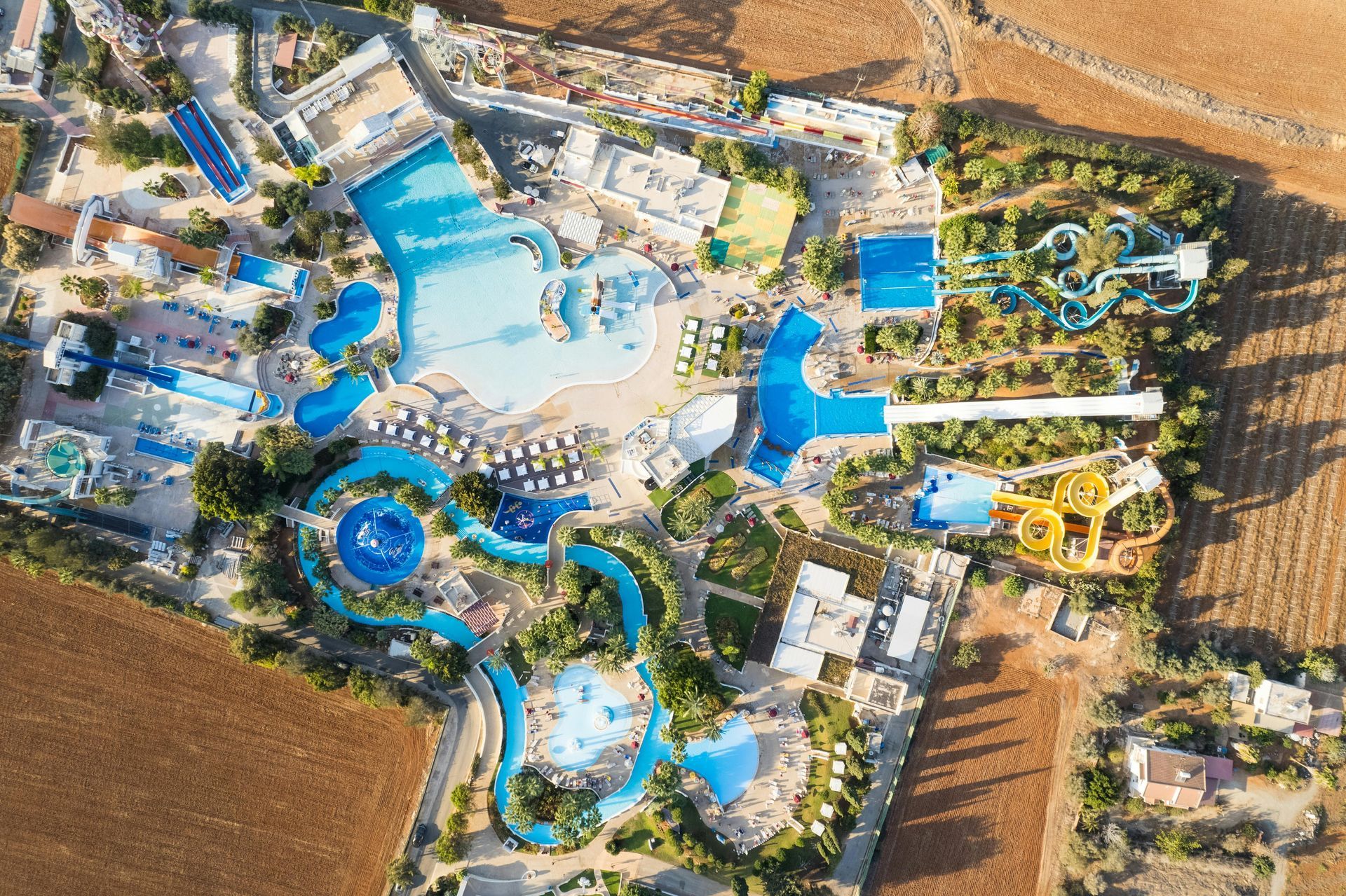 Aerial view of a water park with blue pools, slides, and lounge areas beside a brown field.