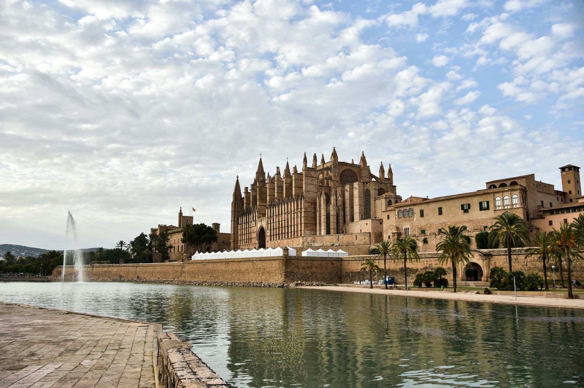 Cathedral beside a reflecting pool under a cloudy sky, with a fountain and historic stone buildings nearby.