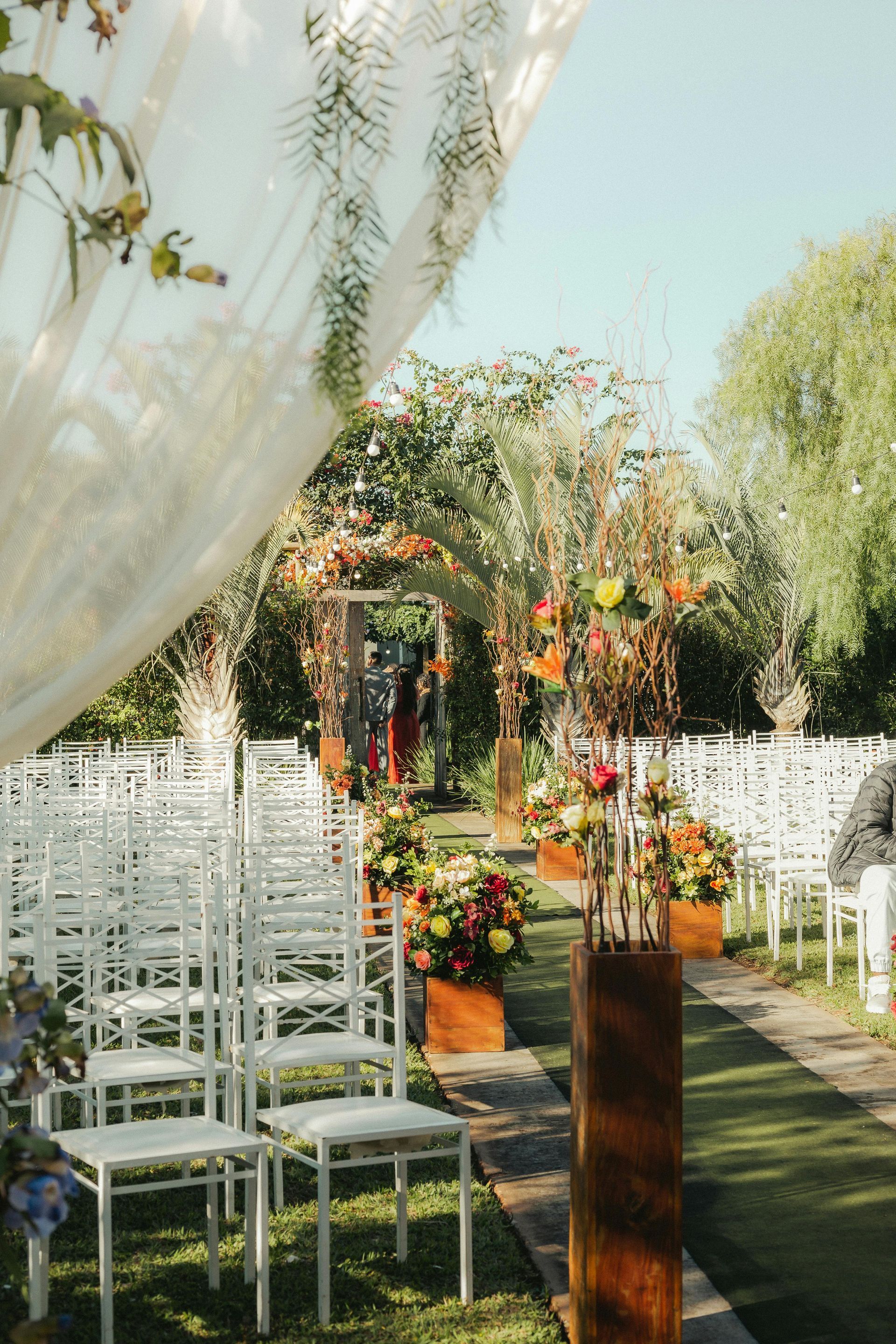 Outdoor wedding aisle with white chairs, floral arch, and draped fabric in a garden setting