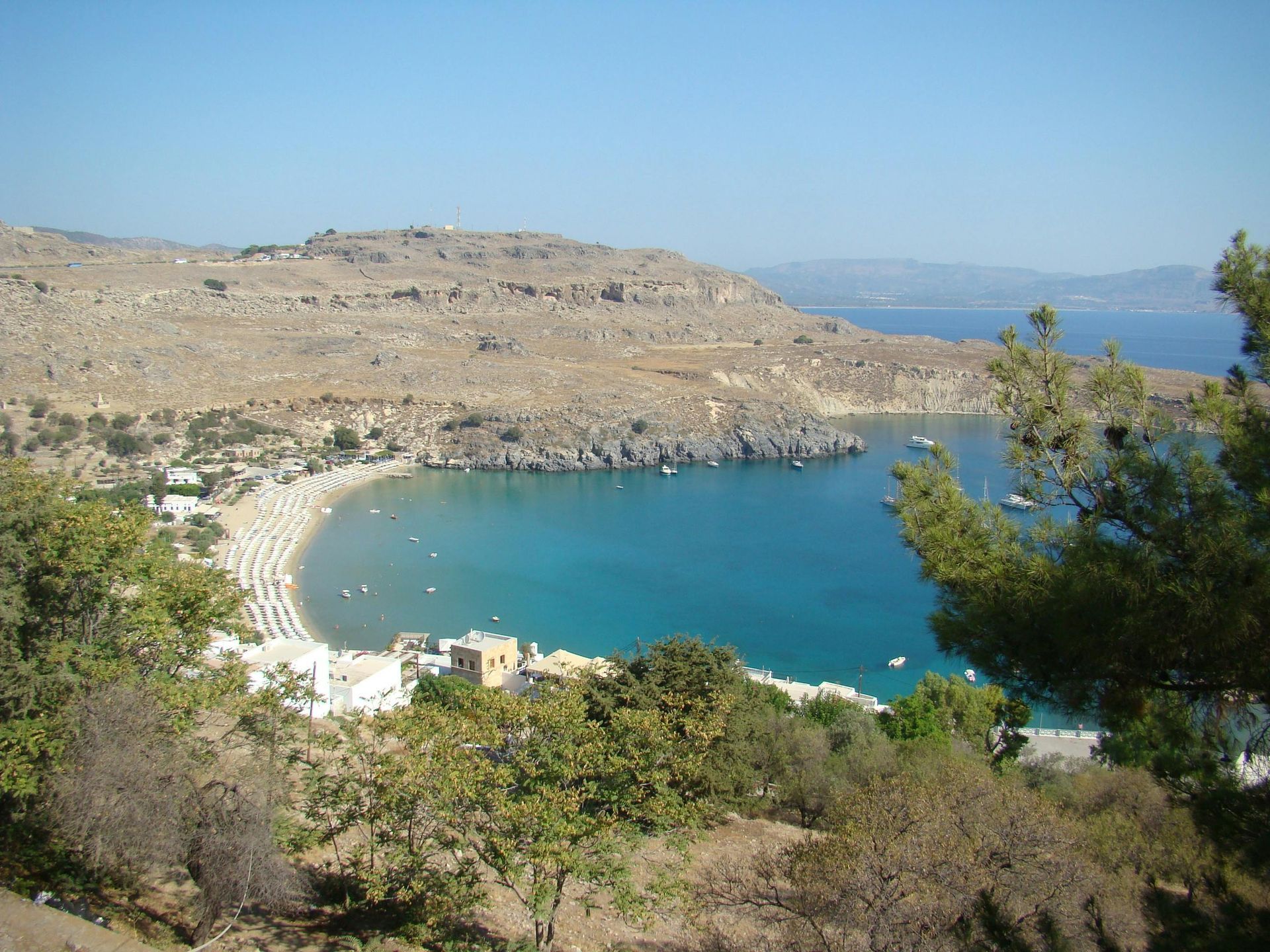 Curved sandy bay with turquoise water, rocky hills, and sparse greenery under a clear sky
