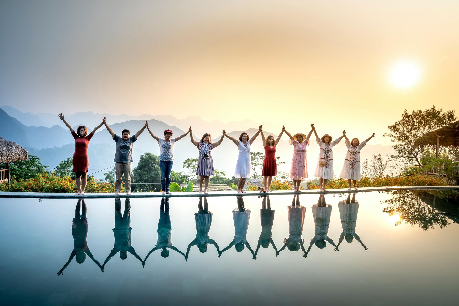 Group of people holding hands on a bridge at sunset, reflected in calm water.