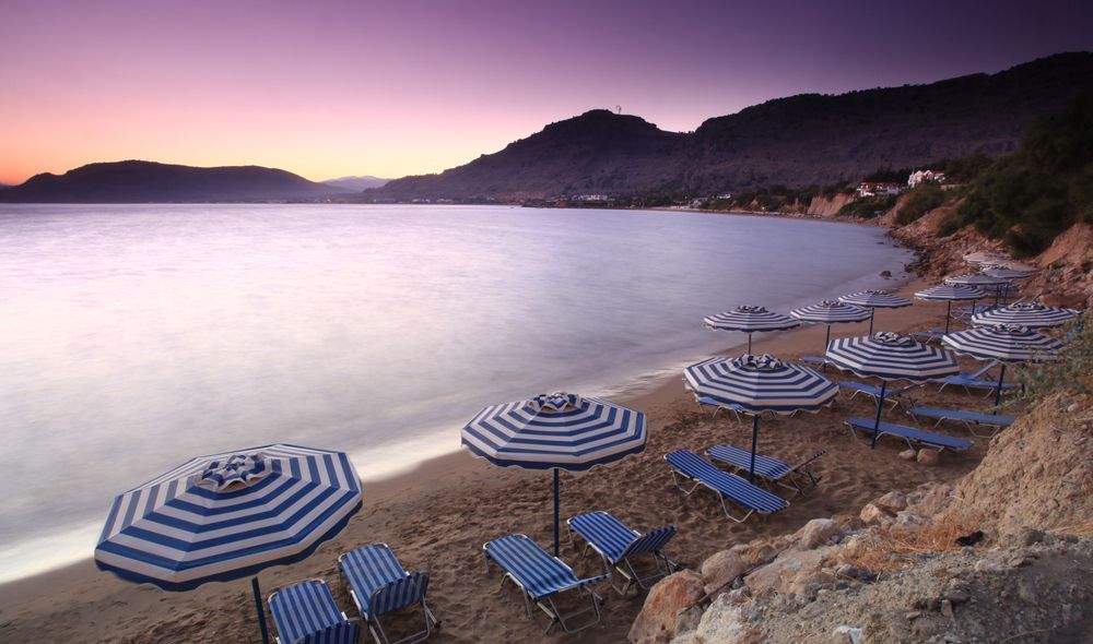 Beach chairs and striped umbrellas on a rocky shore at sunset, overlooking calm water and distant hills