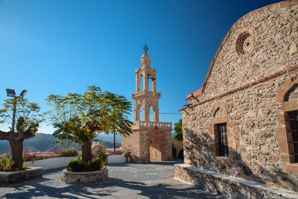 Stone church and bell tower beside a sunlit courtyard under a clear blue sky.